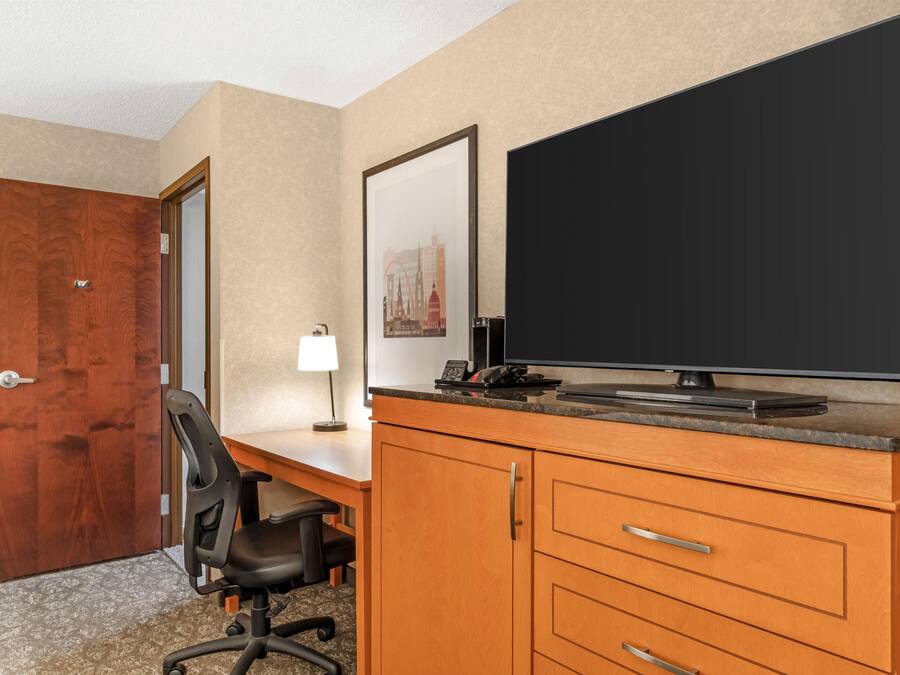 Quarto de hotel com escrivaninha de madeira, cadeira de escritório preta, televisão de tela plana, luminária e porta.