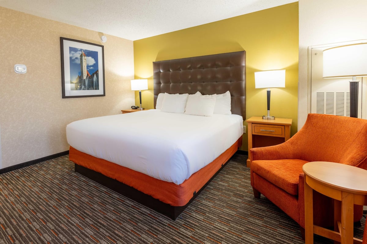 A hotel room with a large bed, two bedside tables with lamps, an orange armchair, and a small wooden table.