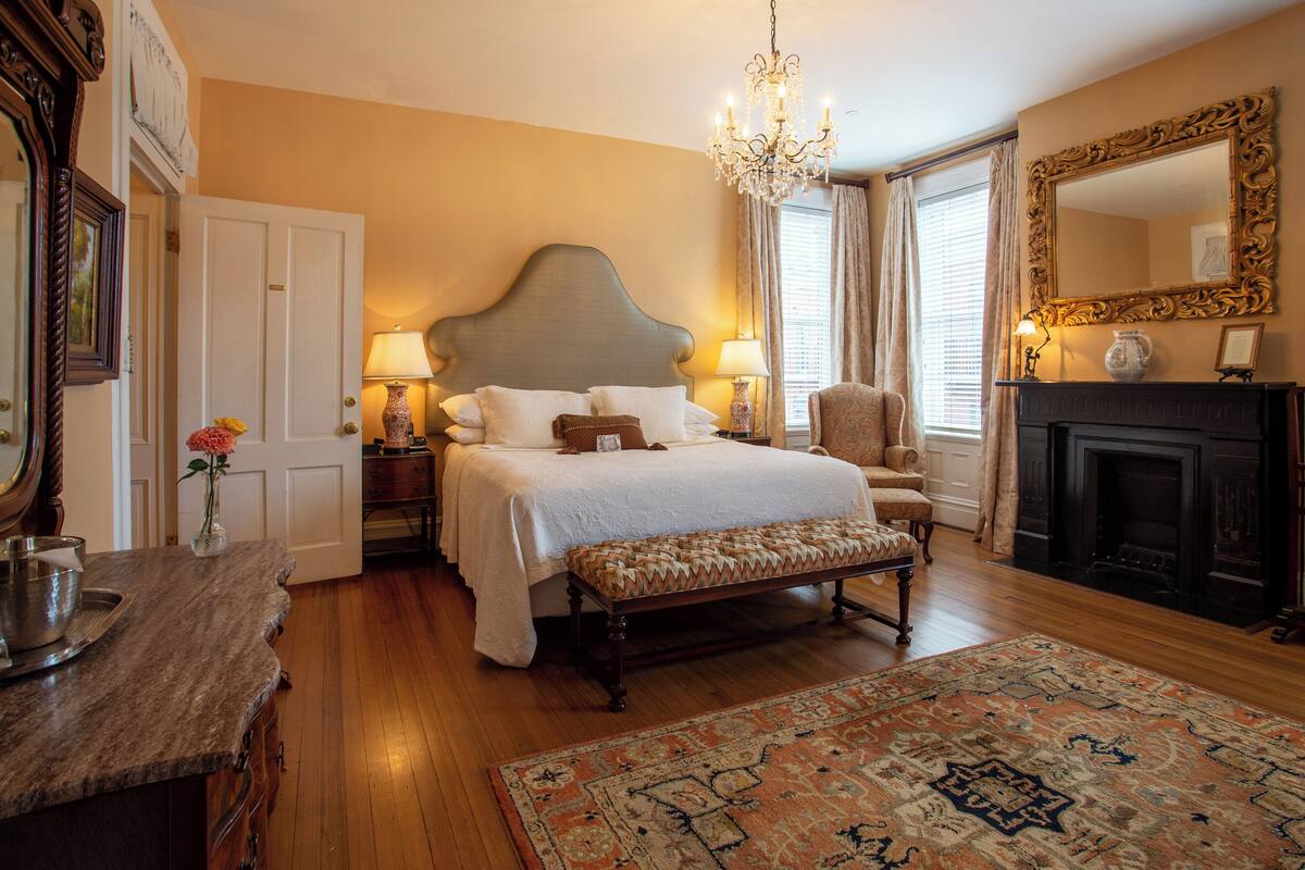 A bedroom with a large bed, a chandelier, a fireplace, and a large mirror.