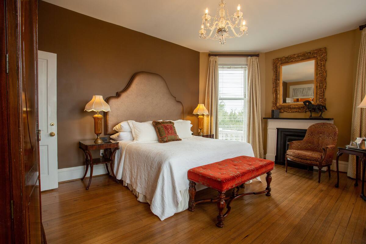 A bedroom with a large bed, two bedside tables with lamps, a chair, a fireplace, and a mirror on the wall.