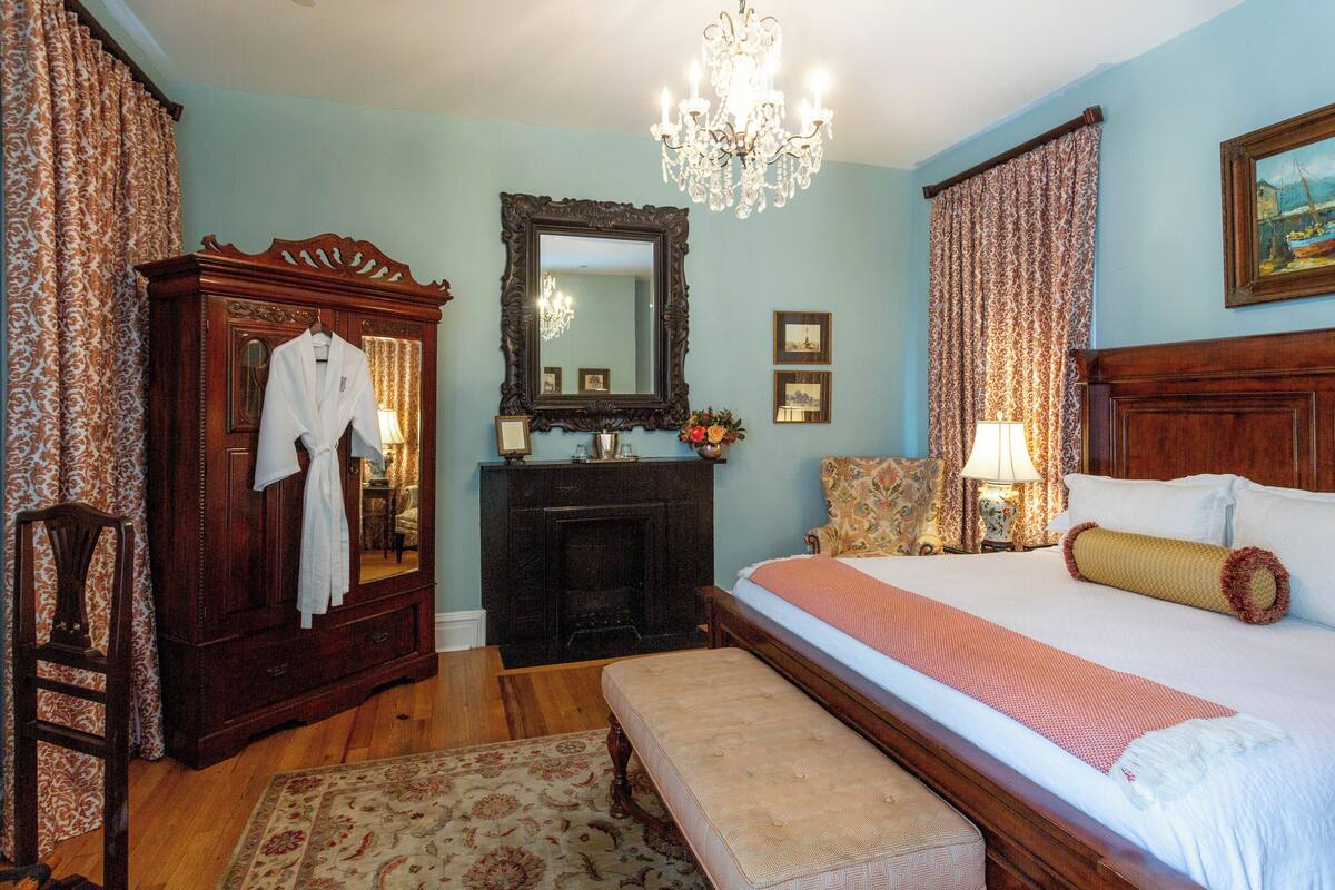 A bedroom with a large bed, a wooden dresser, a mirror, a chandelier, and a fireplace.