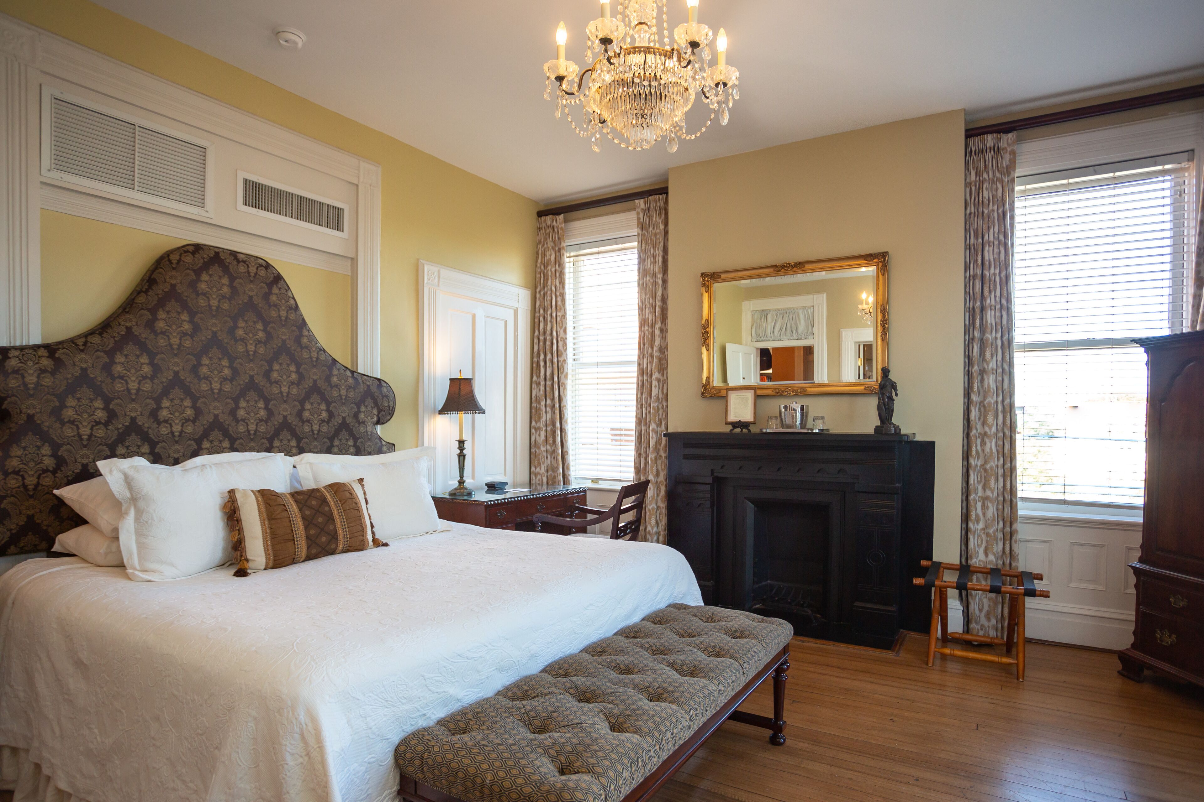 A bedroom with a large bed, a bench, a fireplace, a mirror, and a chandelier.