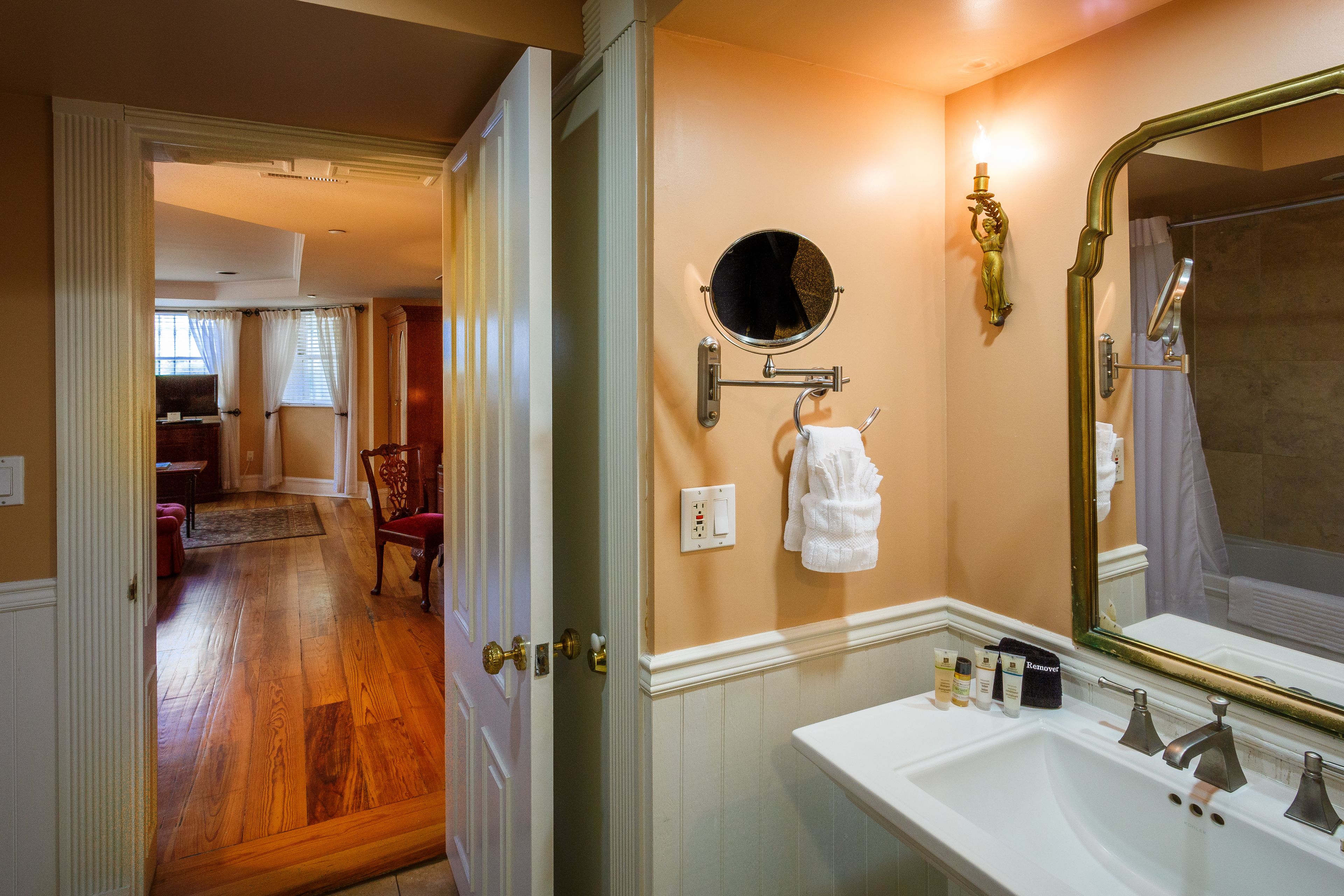 Une salle de bain avec un lavabo blanc, un grand miroir et un miroir rond au-dessus. Une serviette est accrochée au mur, et une petite table contient des articles de toilette.
