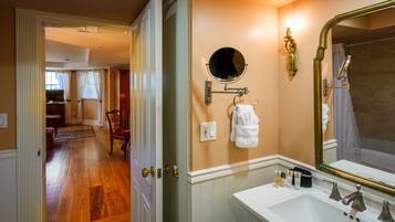 Une salle de bain avec un lavabo blanc, un grand miroir et un miroir rond au-dessus. Une serviette est accrochée au mur, et une petite table contient des articles de toilette.