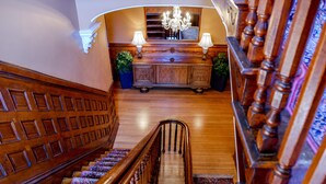 A grand staircase with ornate woodwork, leading to a room with a large wooden cabinet, two lamps, and a chandelier. - Kehoe House, Historic Inns of Savannah (Savannah)
