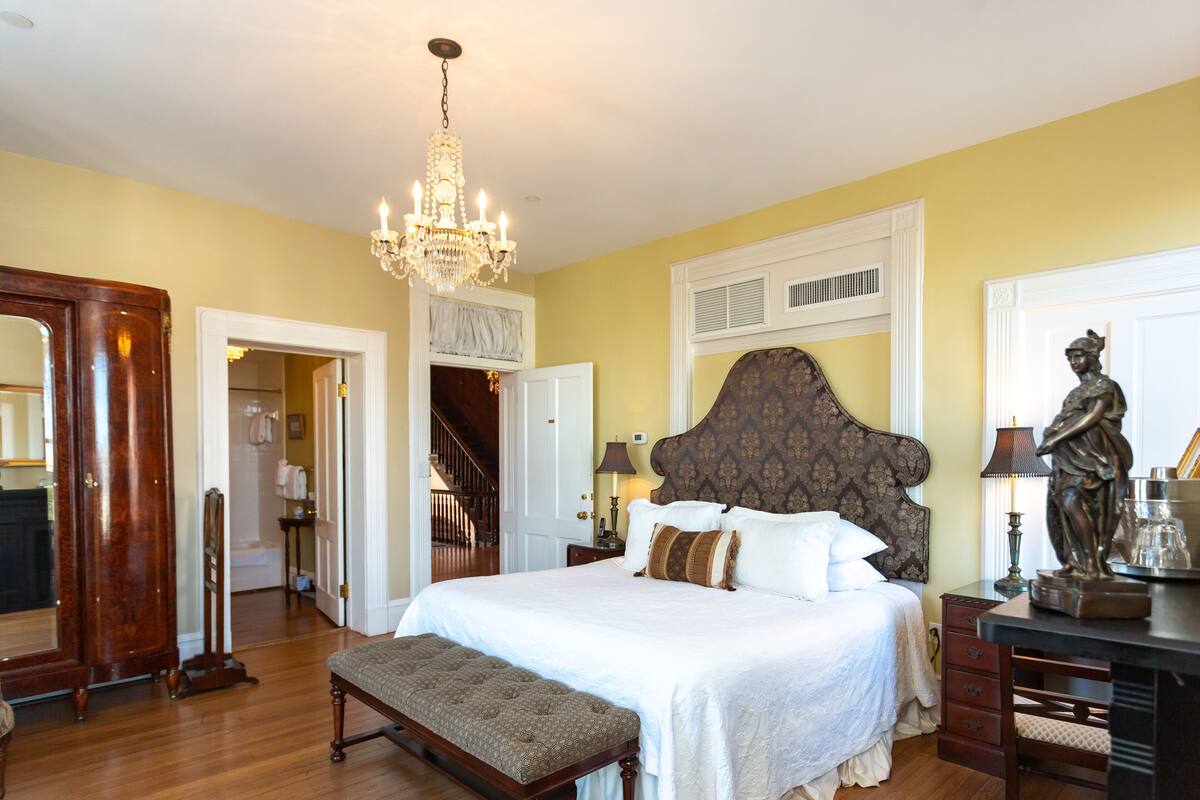 A bedroom with a large bed, a chandelier, a statue, and a wooden bench.