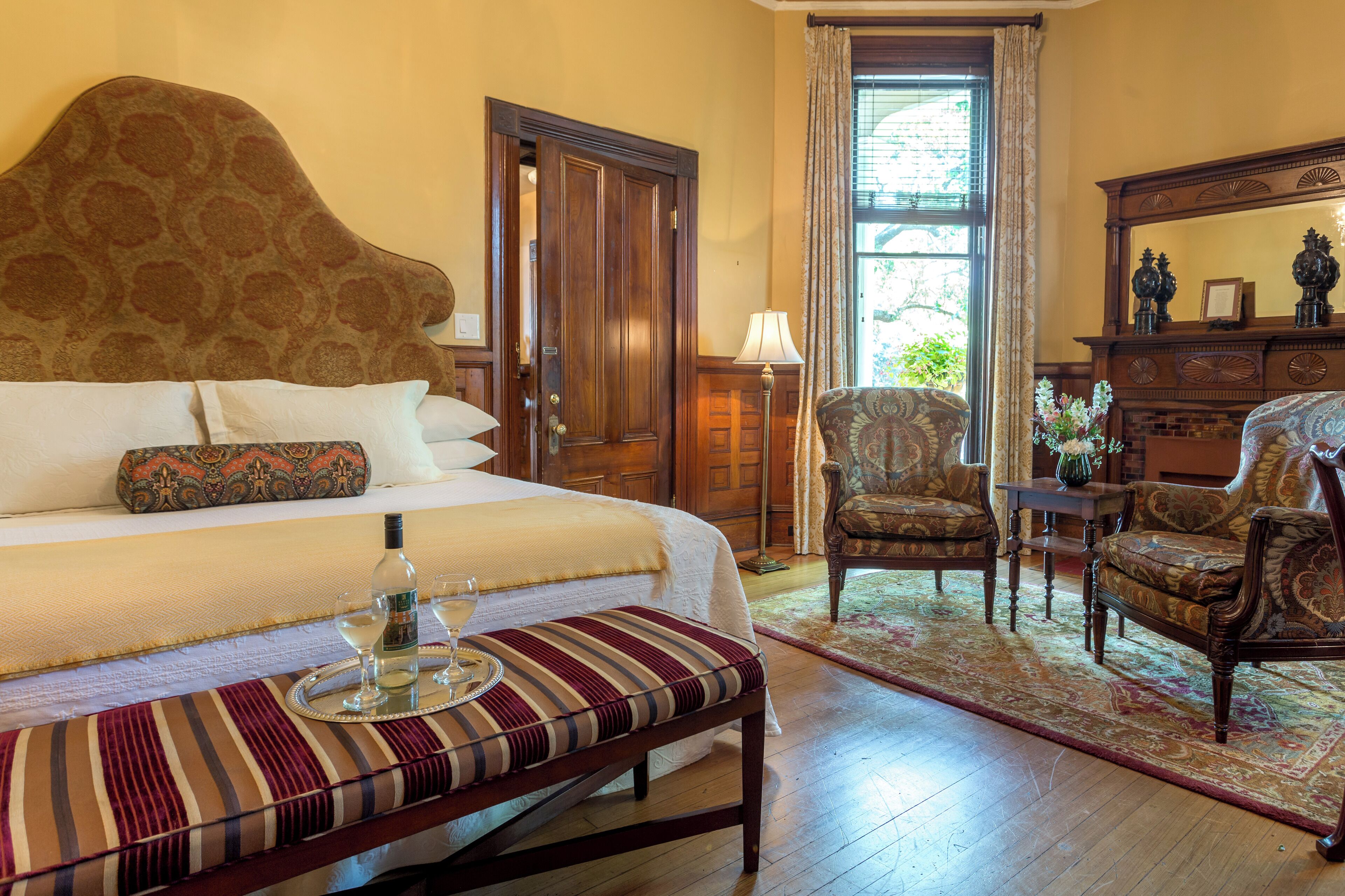 A hotel room with a large bed, a wooden headboard, a bench with a tray, a chair, a table, and a mirror.