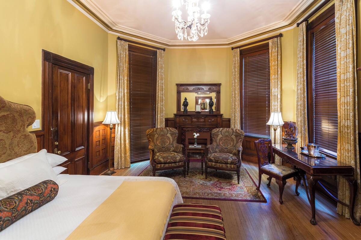 A hotel room with a large bed, two armchairs, a small table, and a mirror above a dresser.