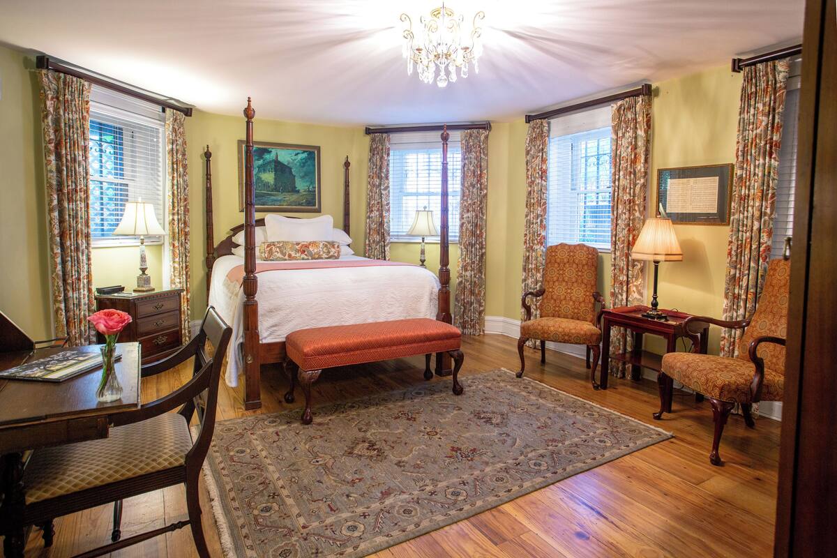 A traditional bedroom with a four-poster bed, a sitting area with a bench and chairs, and a dining table.