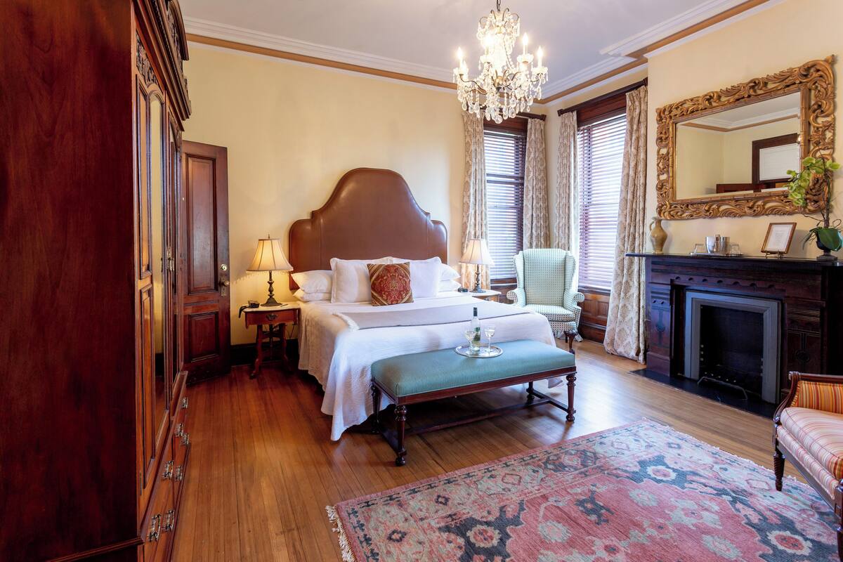 A classic hotel bedroom with a large bed, wooden furniture, a chandelier, and a fireplace.