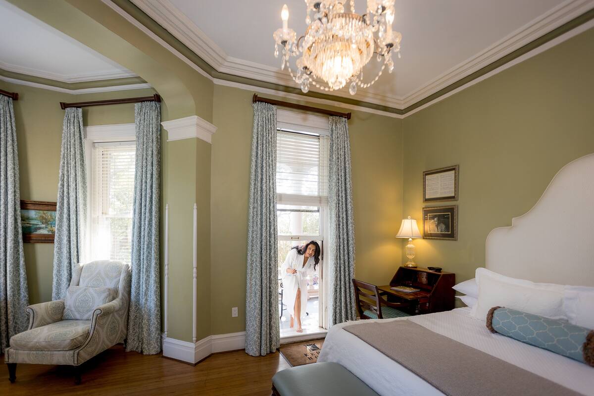A hotel room with a large bed, a sitting area with a chair, a chandelier, and curtains on the windows.