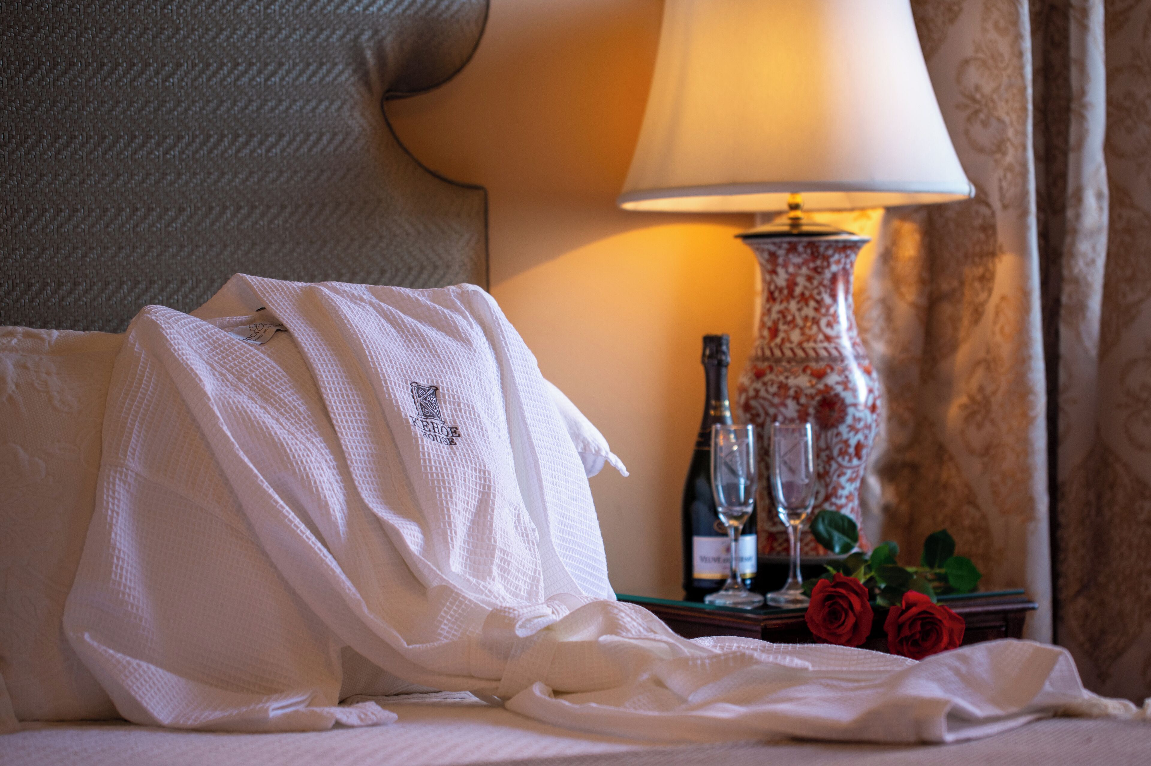 A hotel room with a bed, a white robe, a bottle of champagne, two wine glasses, and a vase with red roses.