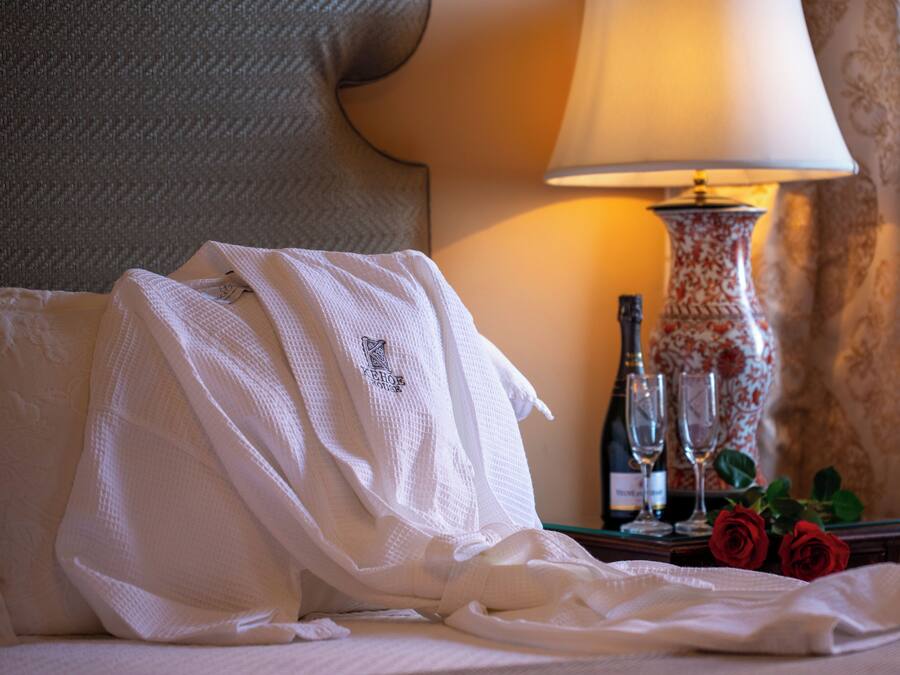 A hotel room with a bed, a white robe, a bottle of champagne, two wine glasses, and a vase with red roses.