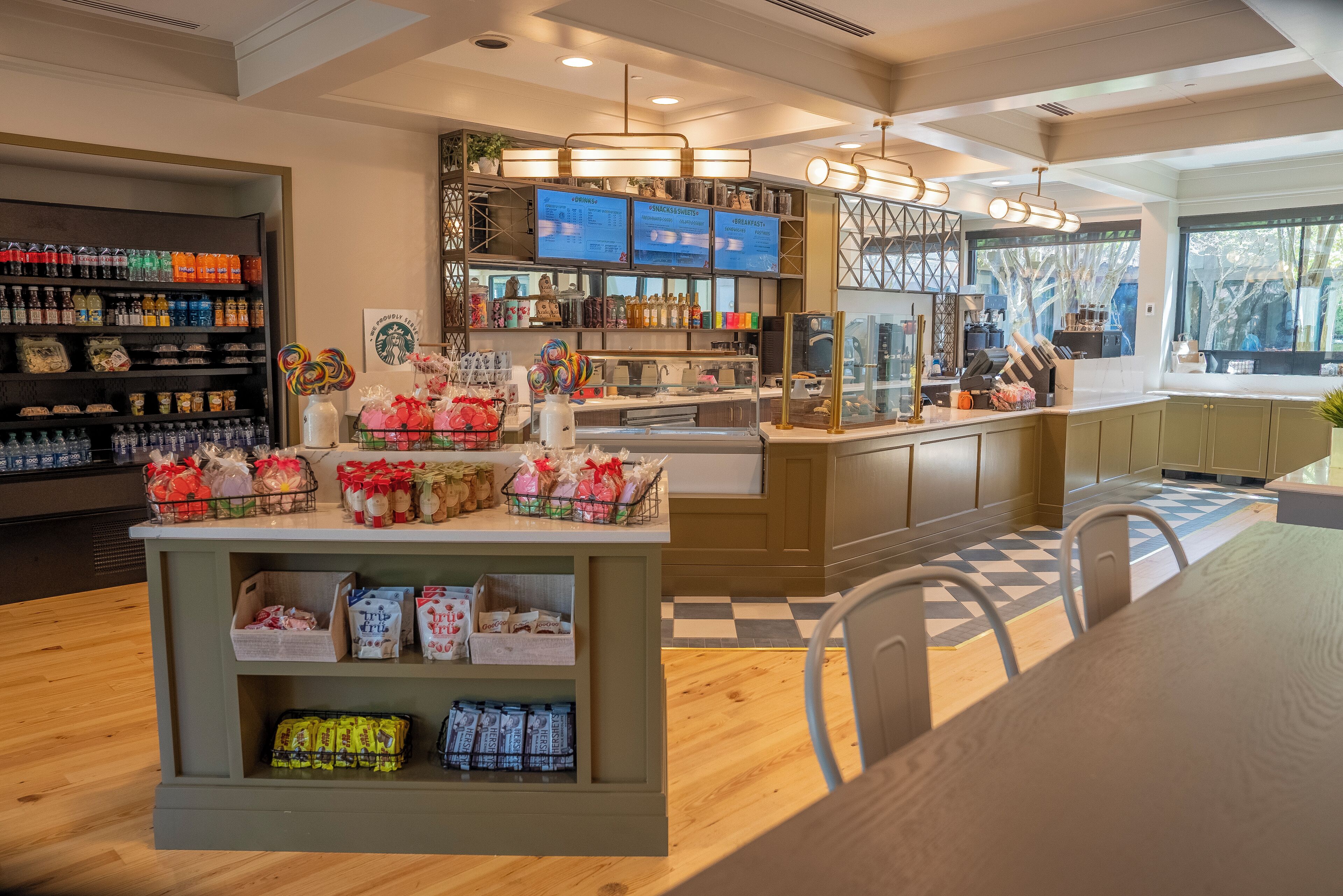 A café interior with a wooden floor, a central counter with a menu board, shelves stocked with bottled beverages, and a seating area with chairs.
