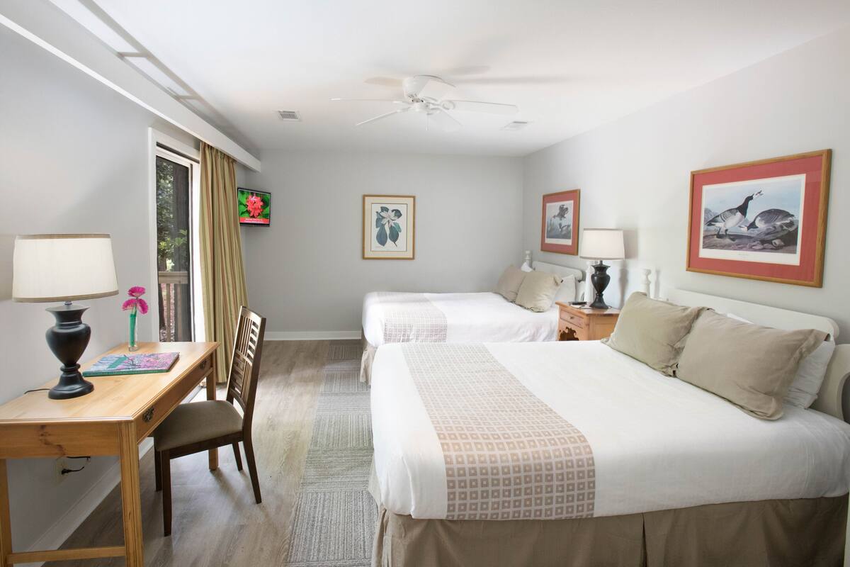A hotel room with two beds, a desk, a chair, a ceiling fan, and artwork on the walls.