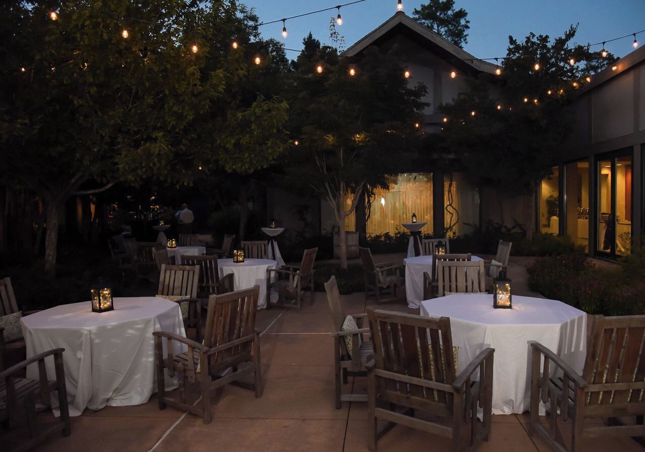 An outdoor dining area with round tables and chairs, string lights, and a building in the background.