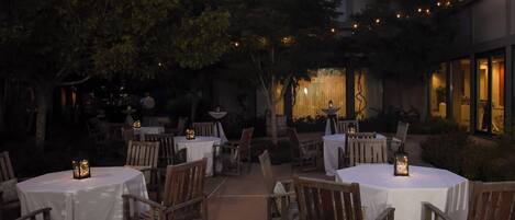 An outdoor dining area with round tables and chairs, string lights, and a building in the background.