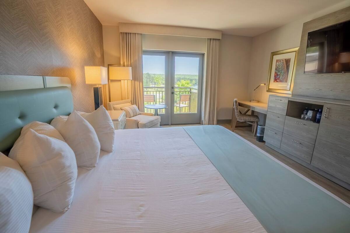 A hotel room with a large bed, a desk, a chair, a balcony with a view, and a TV.