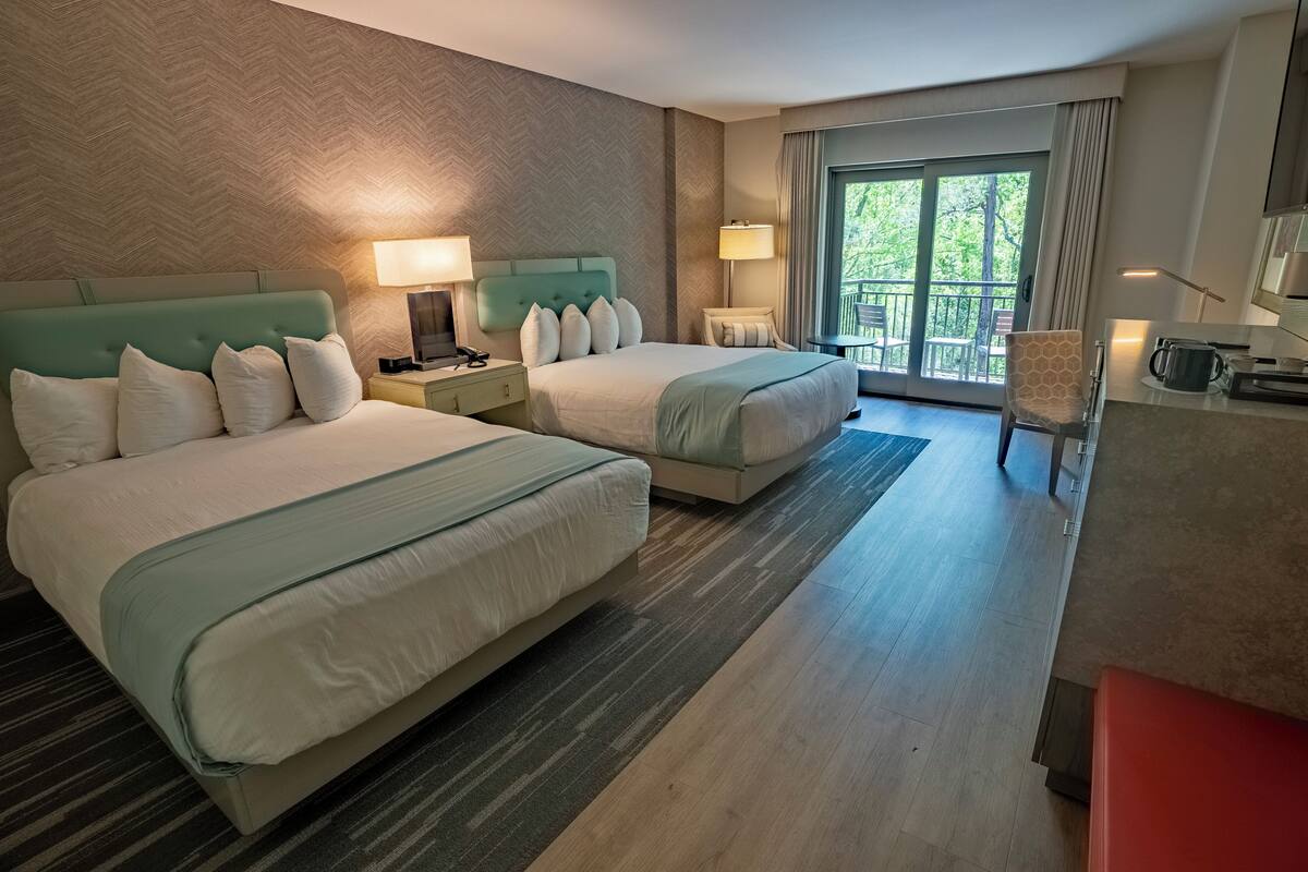 A hotel room with two beds, a desk, a chair, and a balcony with a view of greenery.