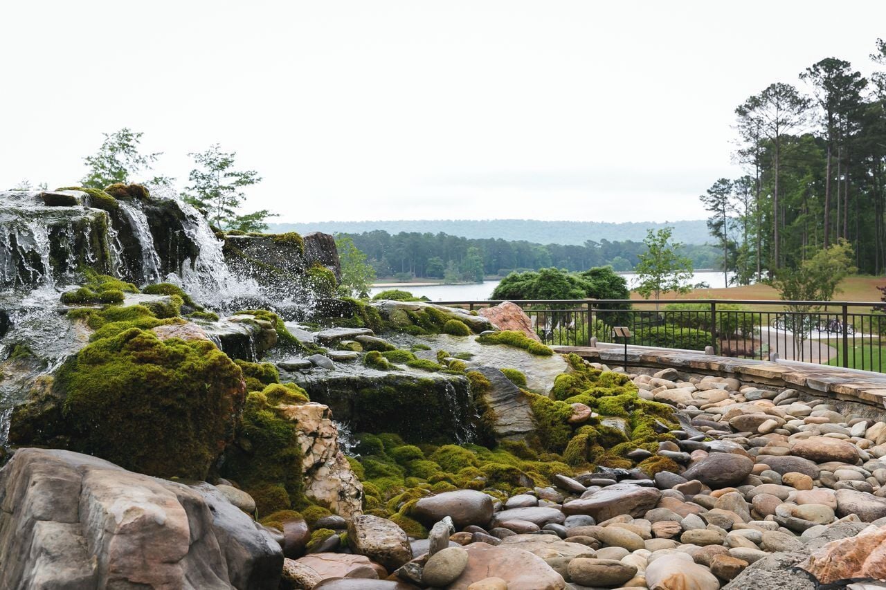 A waterfall cascading over moss-covered rocks with a view of a lake and trees in the background.