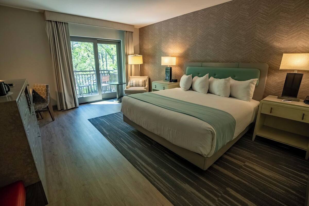 A hotel room with a large bed, two bedside tables with lamps, a chair, a desk with a chair, and a balcony with a view.