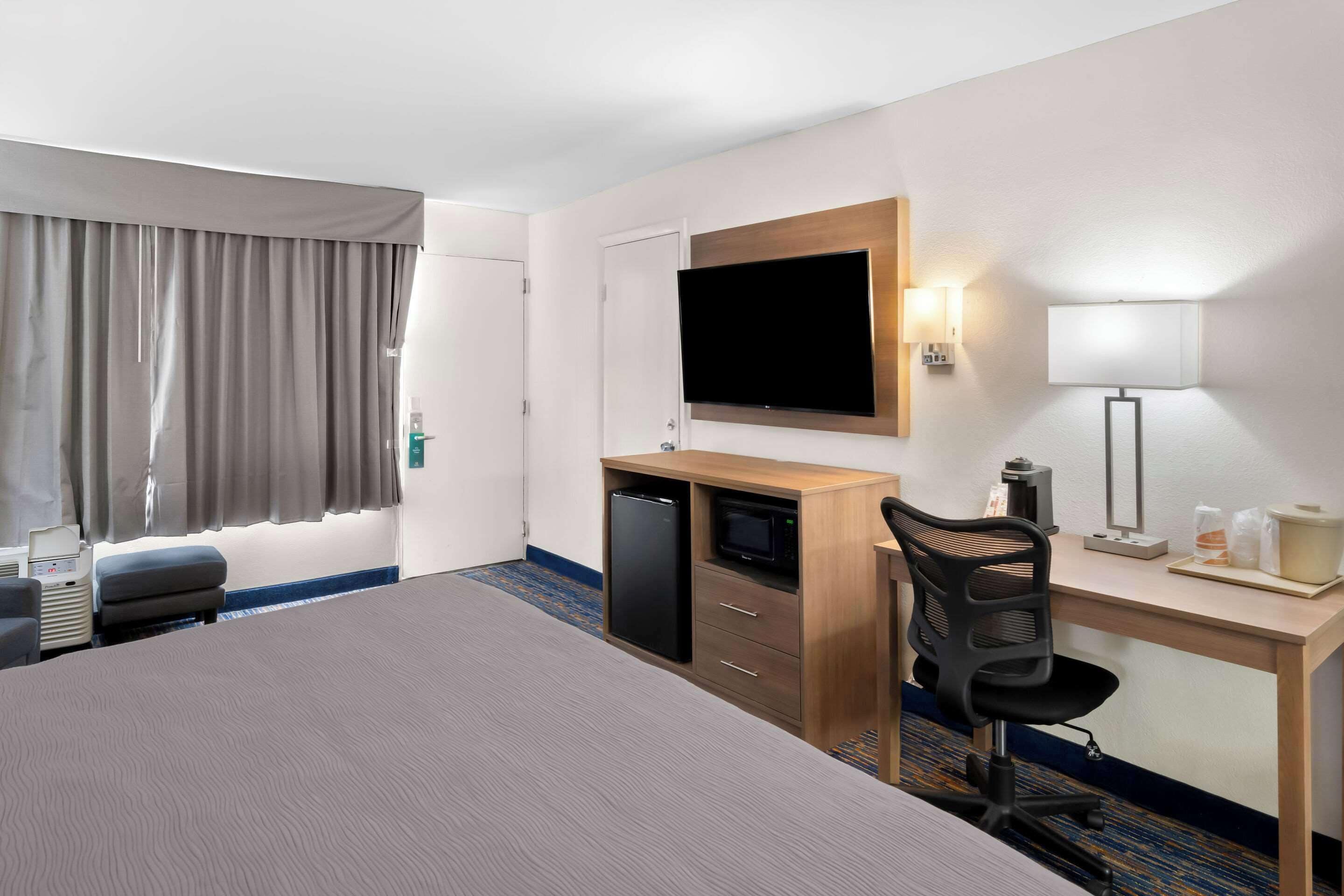 A hotel room with a bed, a desk with a computer, a chair, a television, and a minibar.