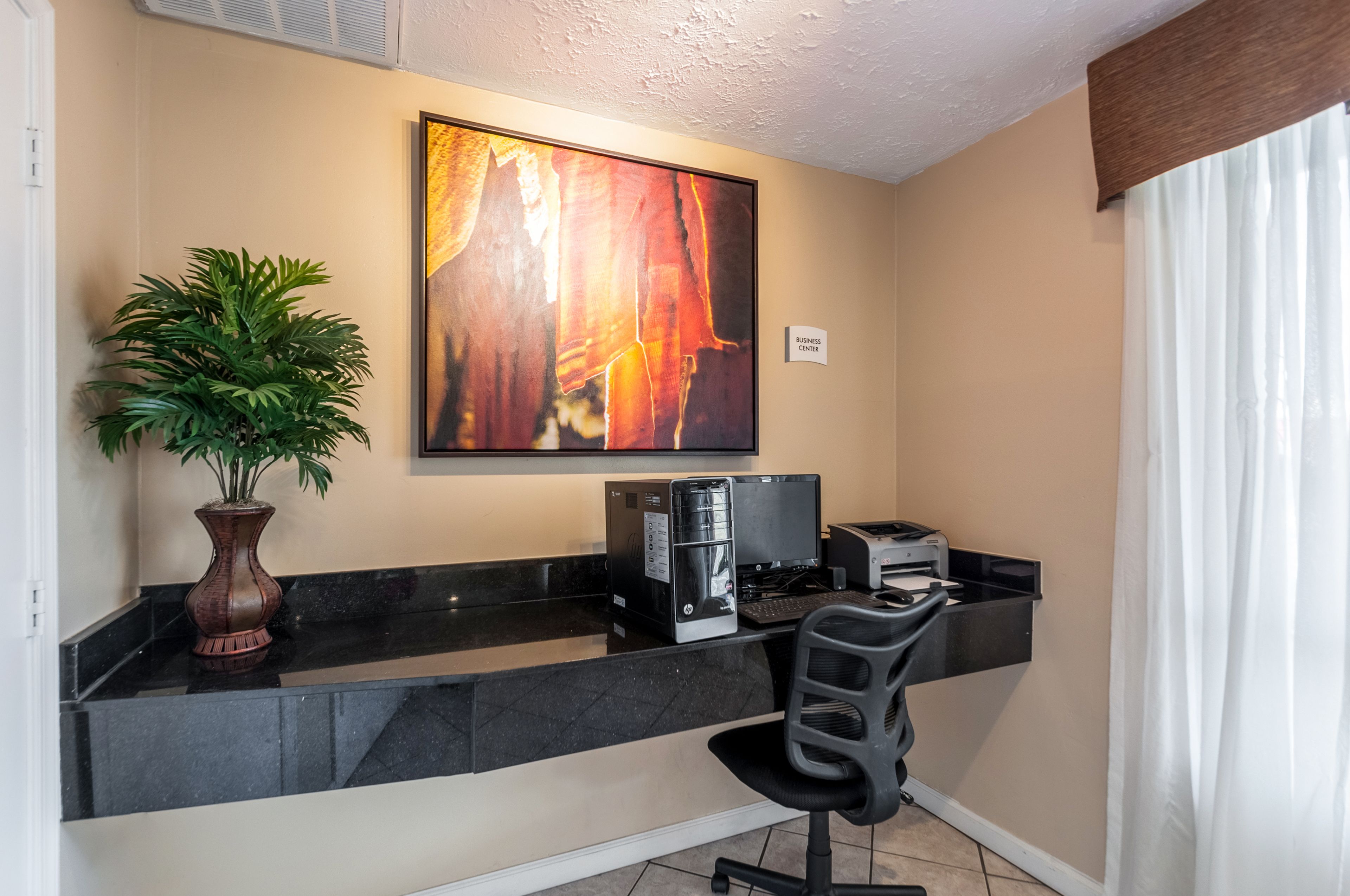 Zona de recepción de hotel con mostrador negro, una máquina de café, una impresora, una silla de escritorio, una planta en maceta y un cuadro abstracto grande en la pared.