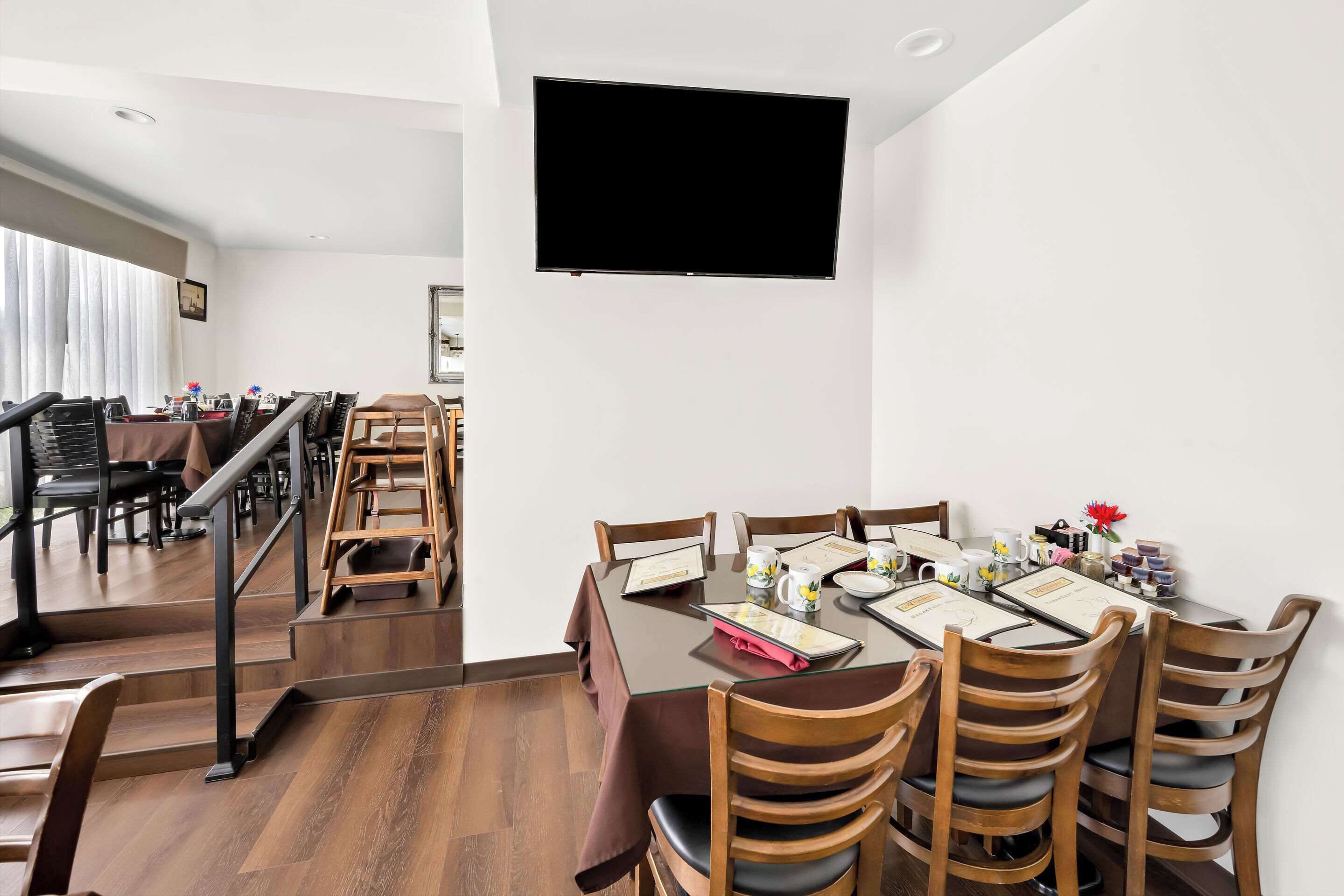 A dining area with a table set for four, wooden chairs, a TV mounted on the wall, and a staircase leading to another room.