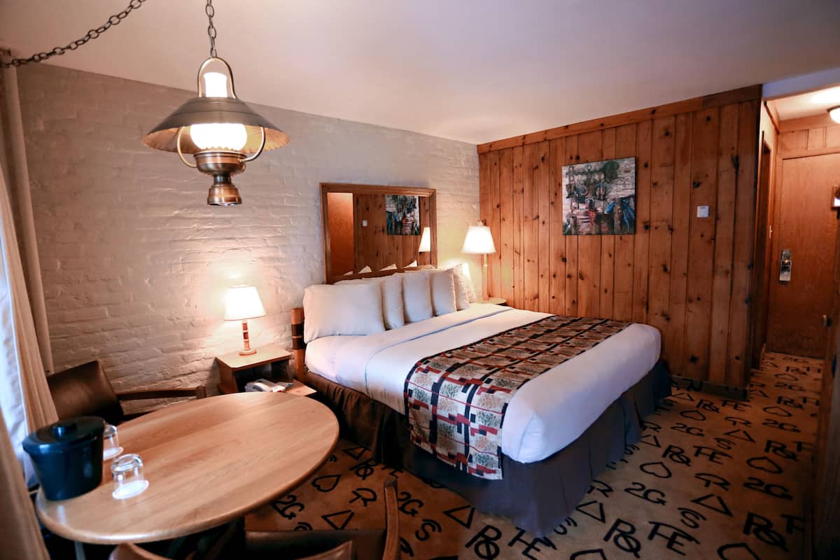 A hotel room with a wooden bed, a round wooden table, a lamp, and a painting on the wall.