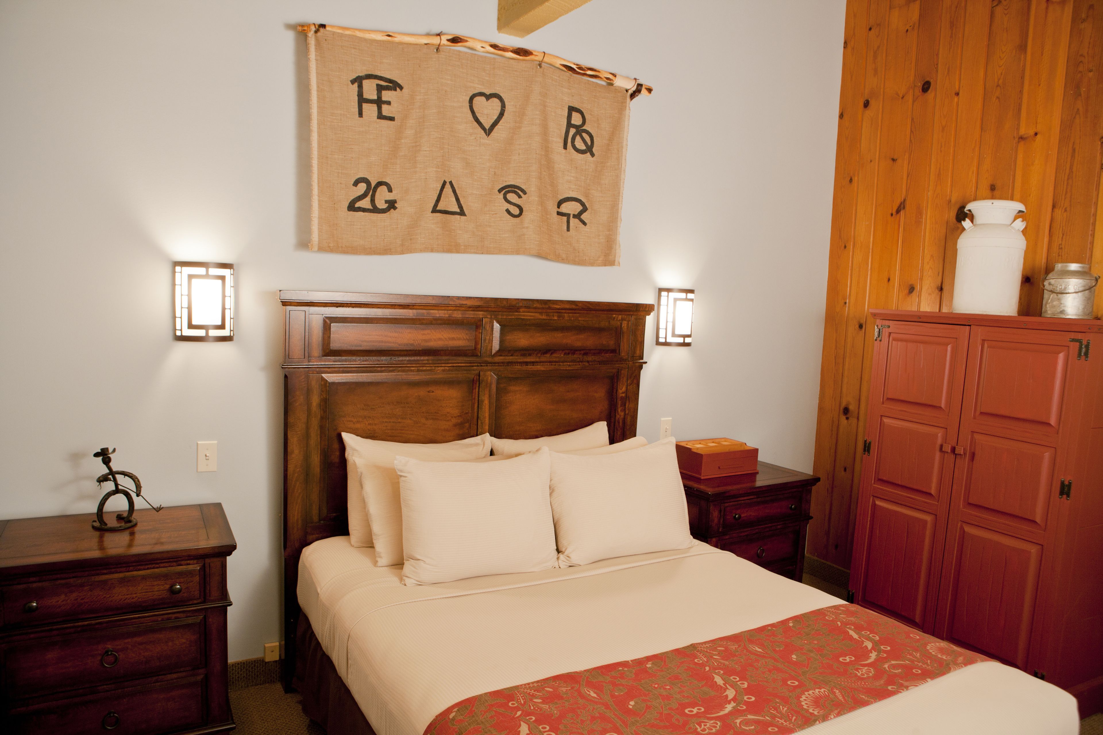 A bedroom with a wooden headboard, a bed with white bedding, a red patterned throw, a wooden nightstand, a small table, and a wooden wardrobe.