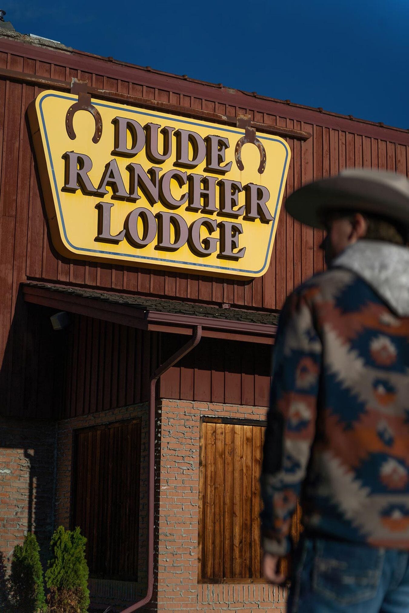 Un letrero que dice 'Dude Rancher Lodge', con fachada de madera y una persona vestida de vaquero.