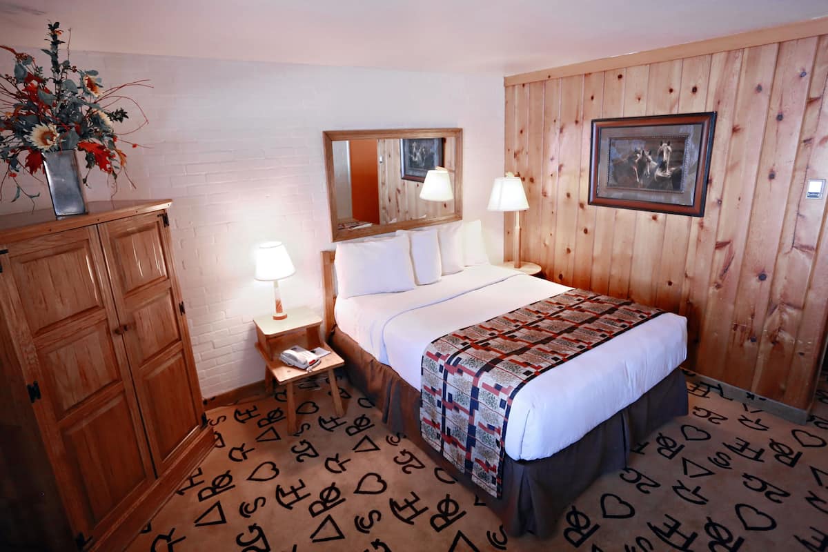 A hotel room with a wooden wardrobe, a bed with a quilt, a bedside table with a lamp, a mirror above the bed, and a picture on the wall.