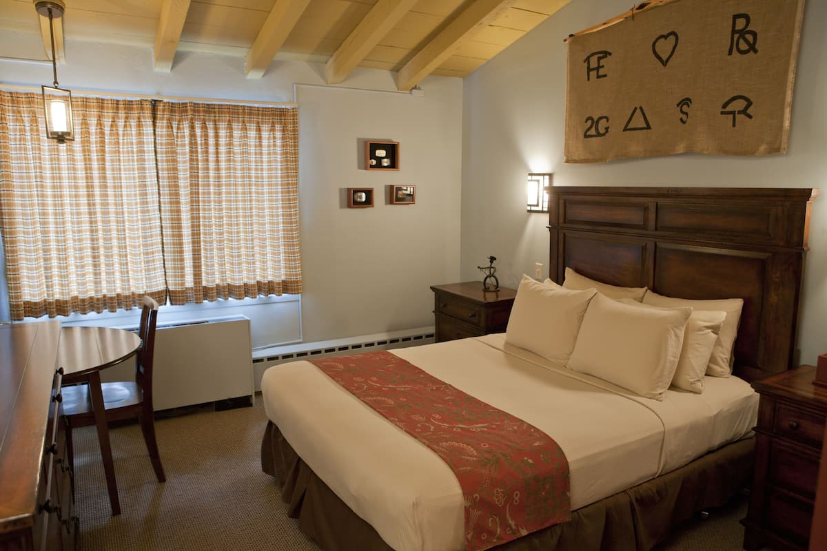 A bedroom with a bed, bedside tables, a nightstand, a window with curtains, a wooden ceiling, and a wall with a hanging sign.