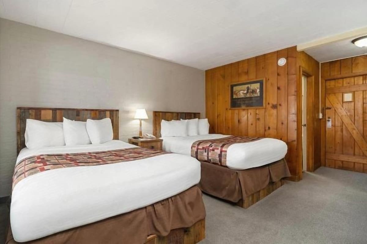 Camera d'albergo con due letti, una parete in legno, una porta e un tavolino con una lampada.