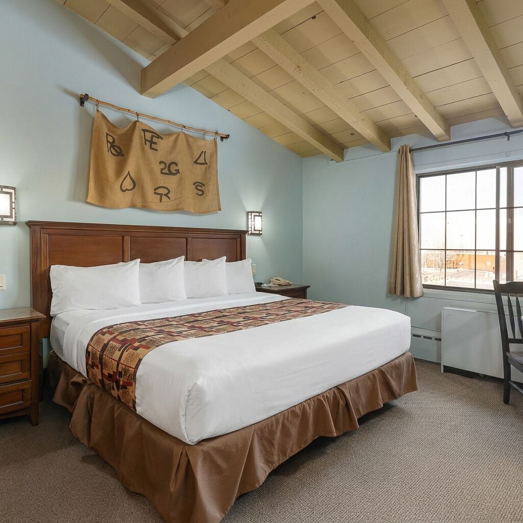 A hotel room with a large bed, wooden headboard, a desk with a chair, a window with curtains, and a wall-mounted sign.