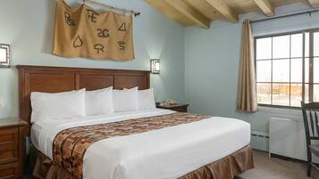 A hotel room with a large bed, wooden headboard, a desk with a chair, a window with curtains, and a wall-mounted sign.