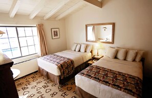 A hotel room with two beds, a window with curtains, a bedside table with a lamp, and a mirror on the wall.