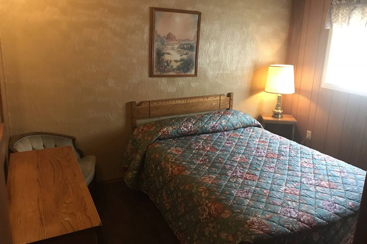 A bedroom with a wooden headboard, a floral bedspread, a nightstand with a lamp, and a painting on the wall.