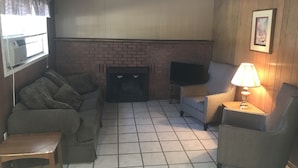 Ein Wohnzimmer mit Kamin aus Ziegelsteinen, einem Fernseher auf einem Schrank, einer Couch, einem Sessel und einem Tisch.