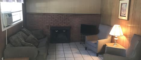 Un salon avec une cheminée en briques, un téléviseur posé sur un meuble, un canapé, un fauteuil et une table.