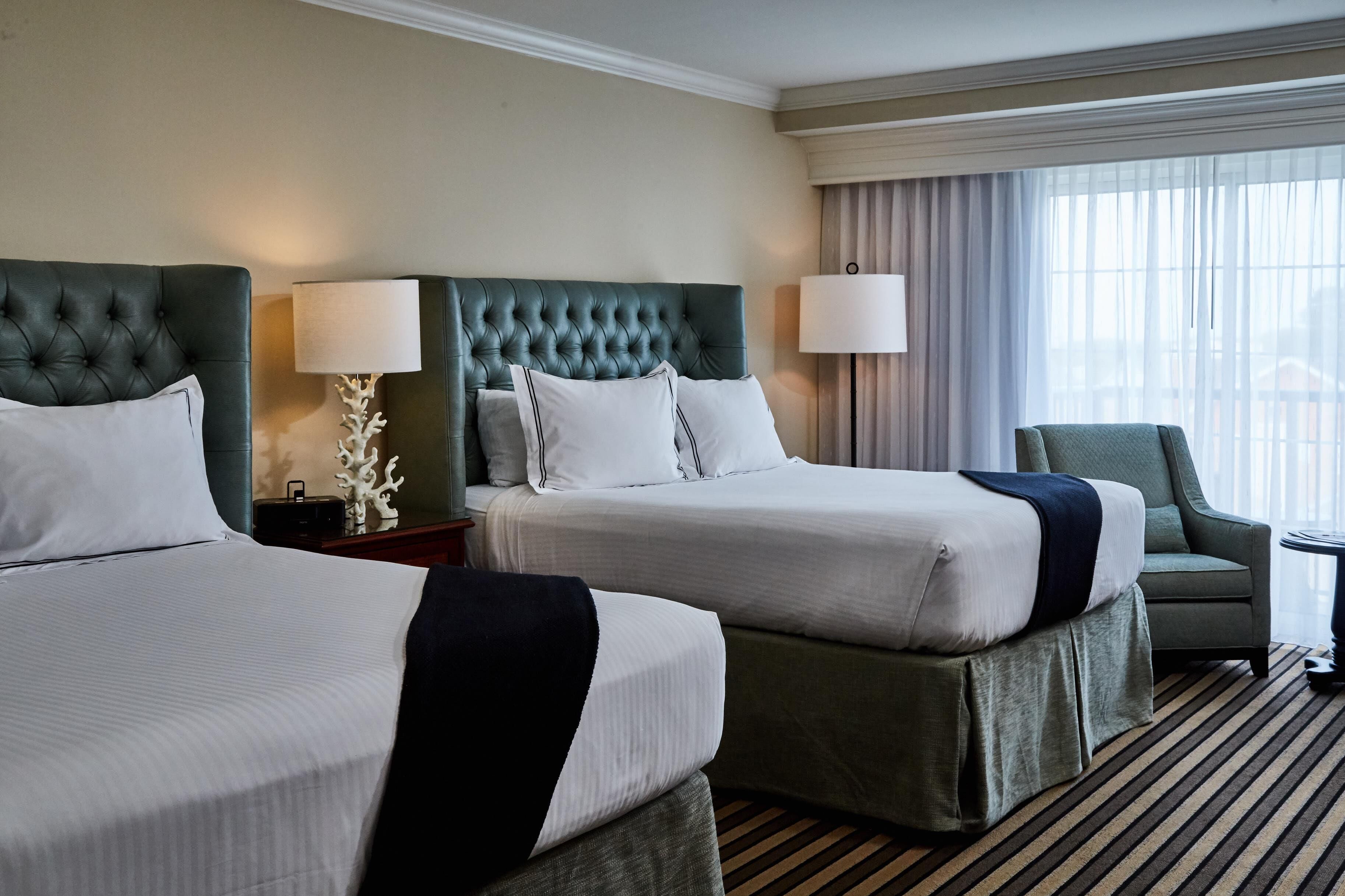 A hotel room with two beds, a green armchair, a small table, and a large window with curtains.