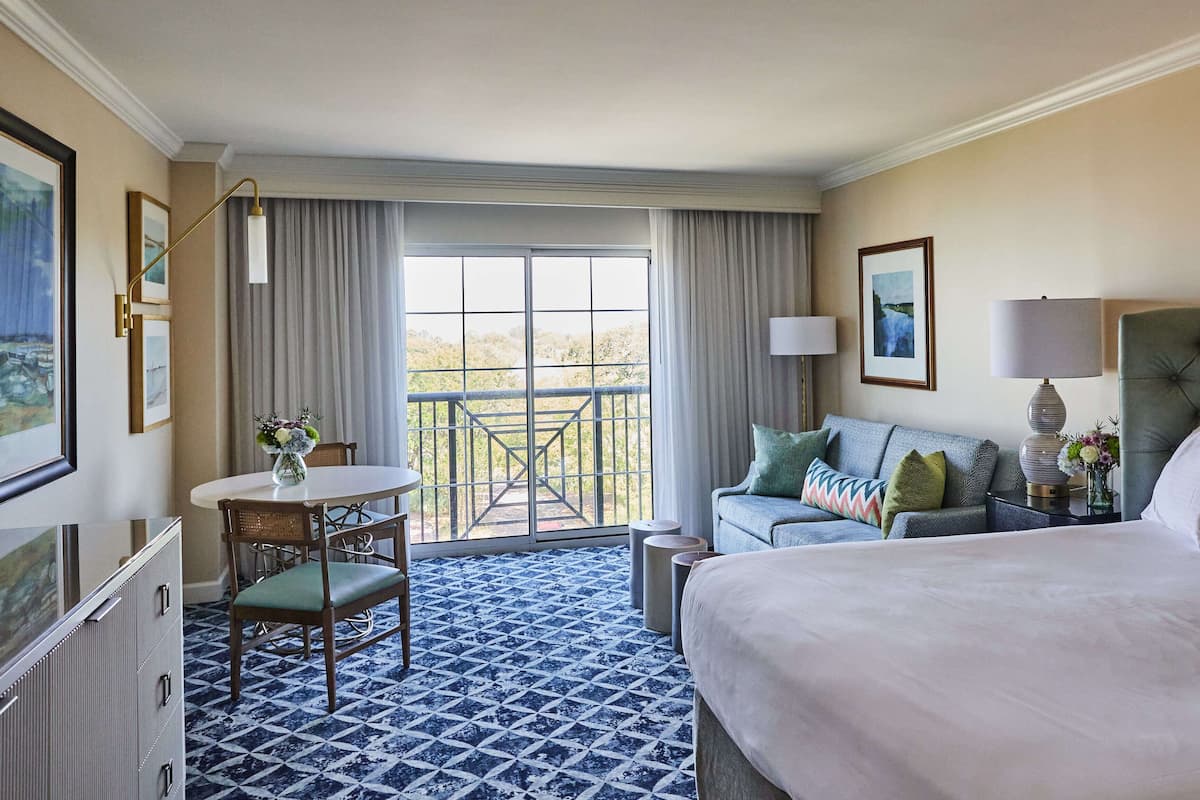 A hotel room with a bed, a sofa, a dining table with chairs, a balcony with a railing, and a view of the outdoors.