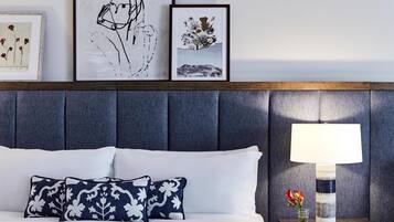 A neatly made bed with white linens and two decorative pillows, a bedside table with a lamp and a vase of flowers, and two framed artworks above the bed.