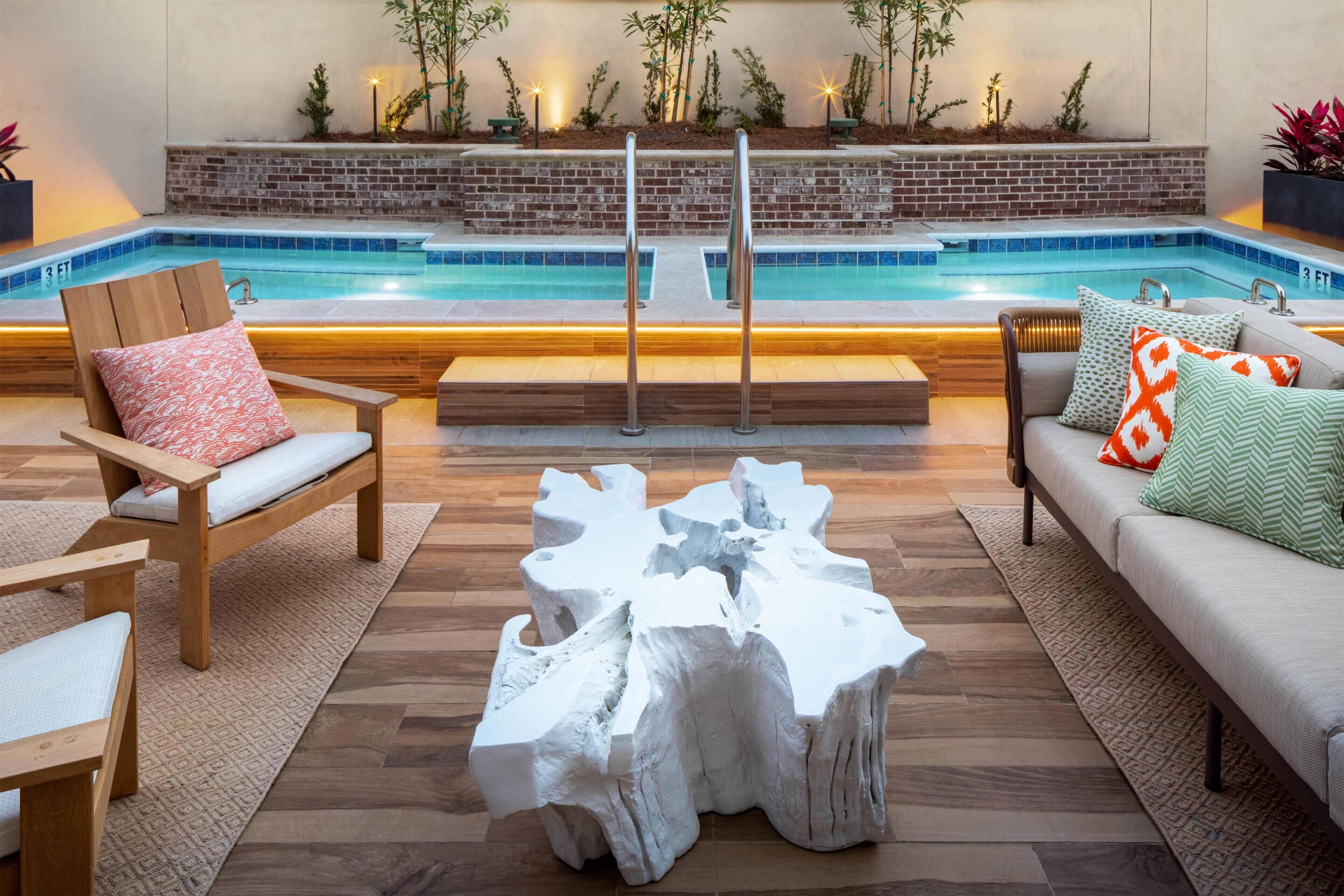 Uma área à beira da piscina com um banco de madeira, uma mesa de centro branca e escultural, e um sofá com almofadas estampadas.