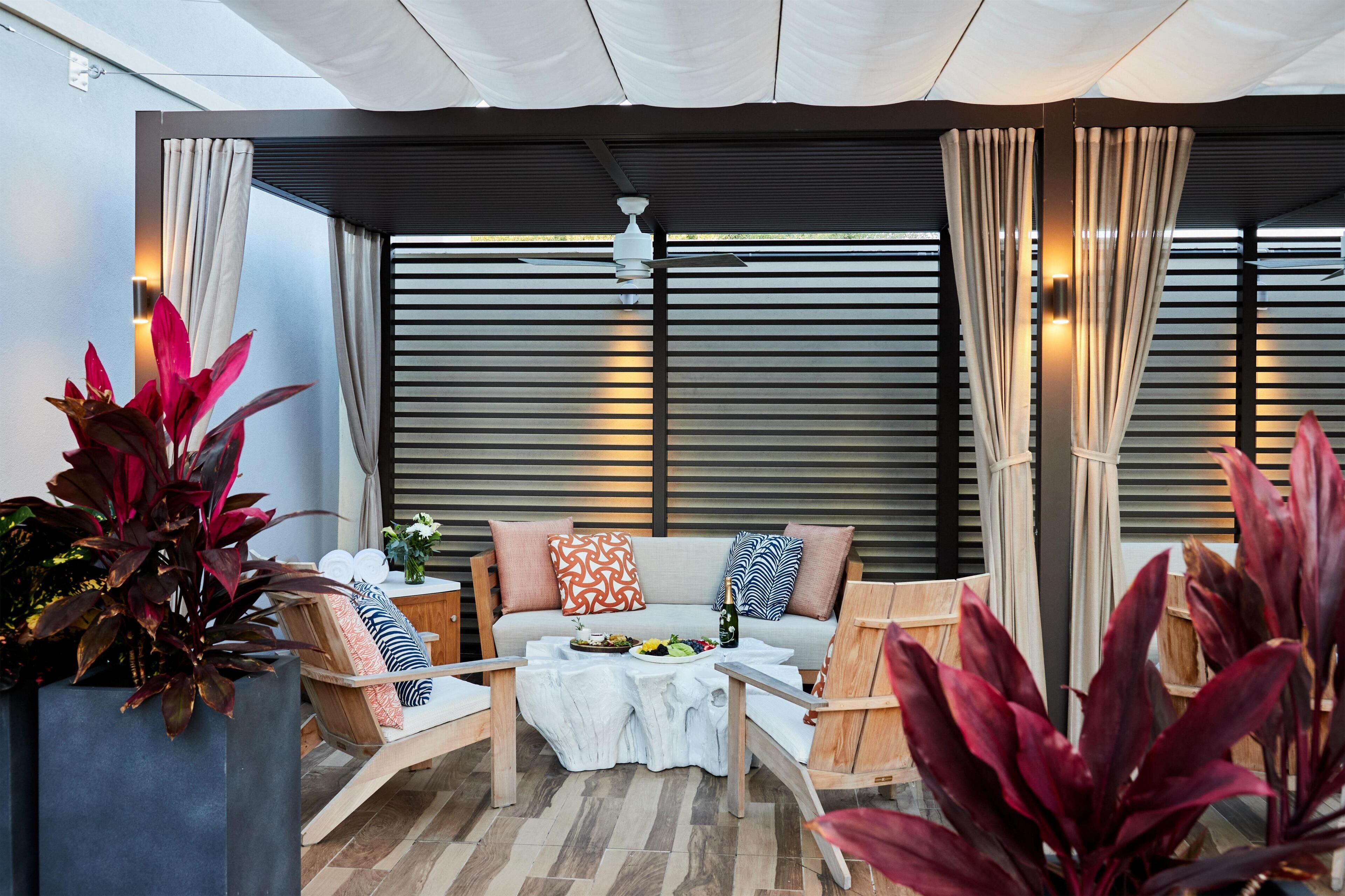 A modern outdoor seating area with a sofa, chairs, and a table, surrounded by large windows and curtains.