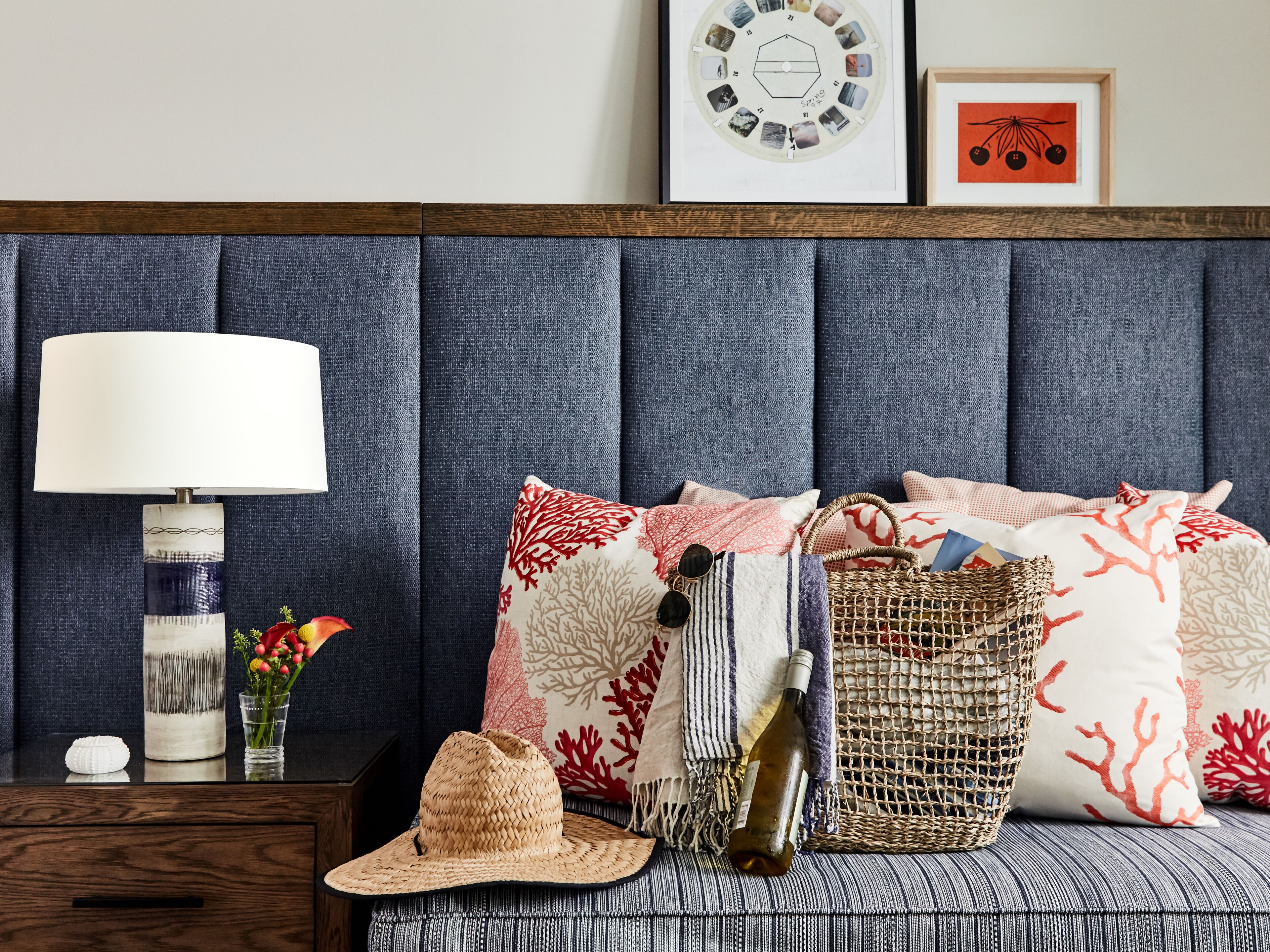 Un dormitorio ordenado con una tapizada en azul, una lámpara de noche, cojines decorativos, una cesta tejida y una botella de vino.