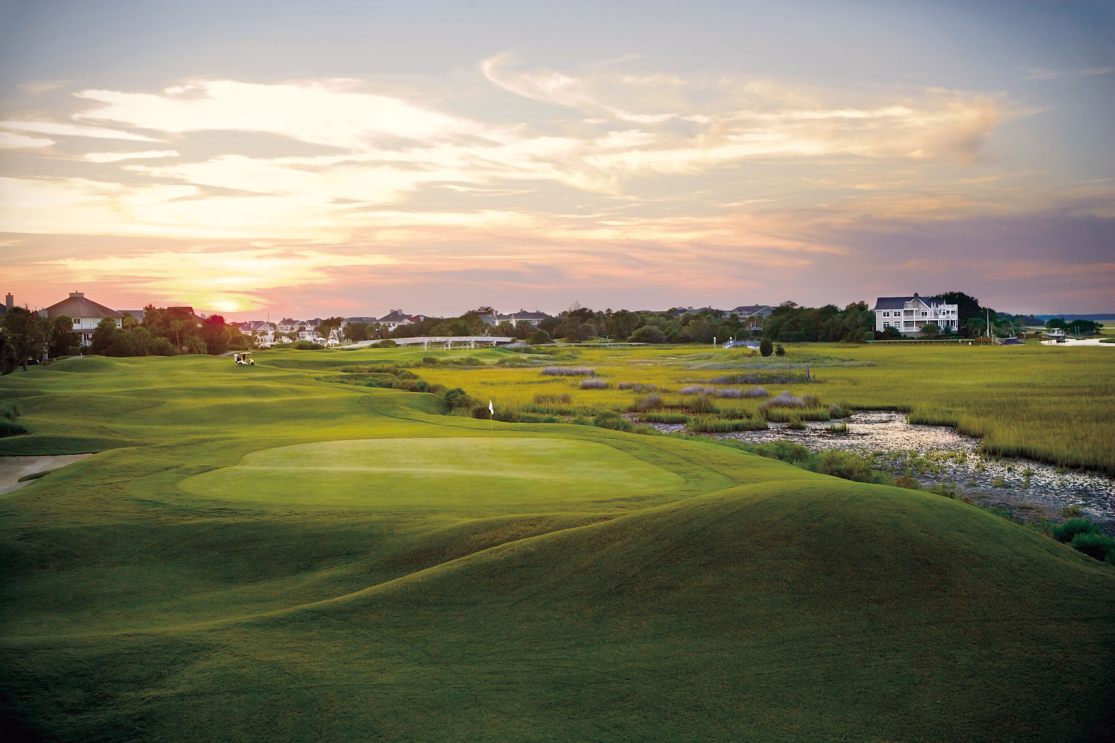 Un parcours de golf avec des collines verdoyantes, un club-house et un coucher de soleil en toile de fond.