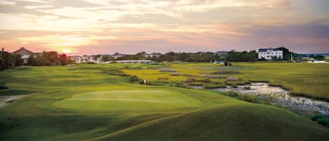 A golf course with rolling green hills, a clubhouse, and a sunset in the background.