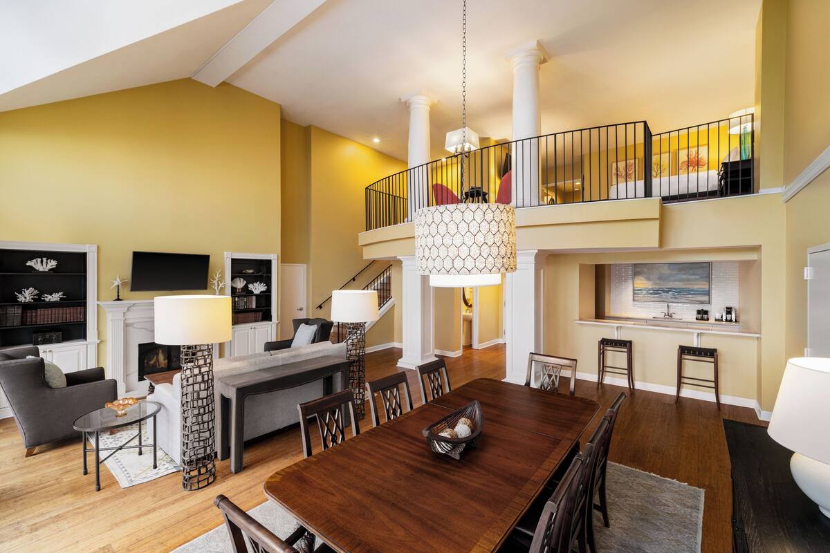 A spacious living room with a dining area, a staircase leading to a balcony, and a kitchen area.