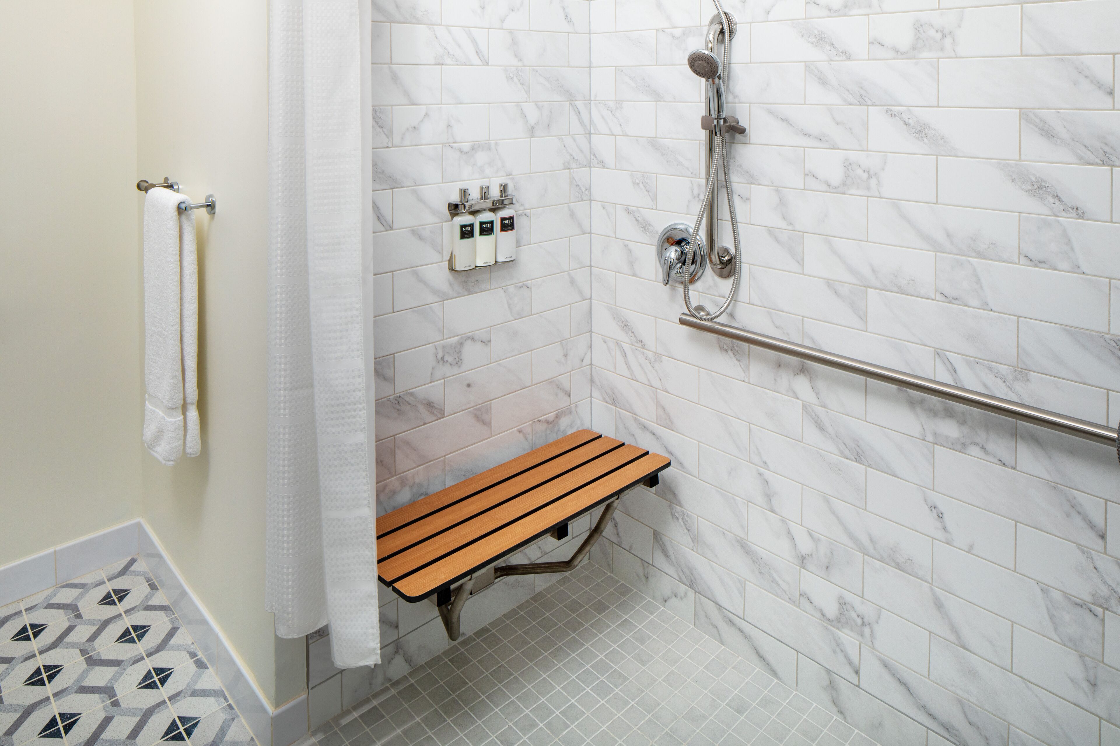 A bathroom with white marble tiles, a wooden bench, a towel rack, a shower head, and a handrail.
